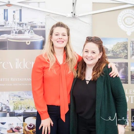 Two women smiling, arms around each other, in front of a promotional display.