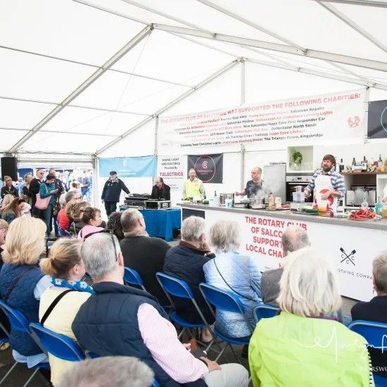 People watch a cooking demonstration under a tent. A chef cooks behind a counter.
