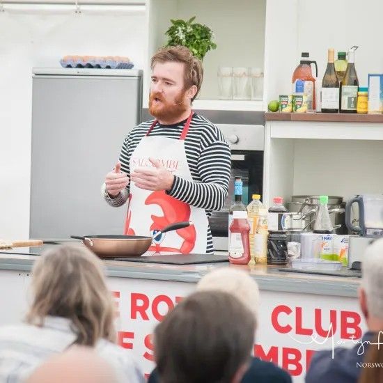 Man in apron cooks in front of audience. Kitchen setting, ingredients on counter, wearing striped shirt, gesturing.