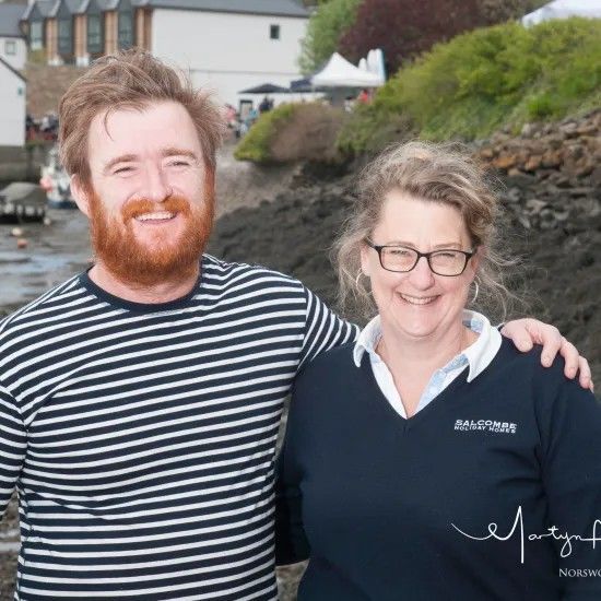Smiling man with a red beard in stripes, arm around smiling woman in glasses, waterfront in background.