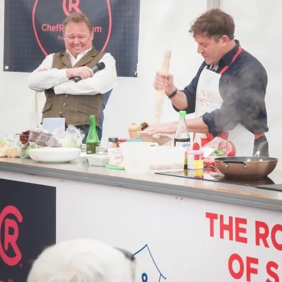 Two chefs cooking on stage; one stirs, the other prepares ingredients. White tent backdrop.