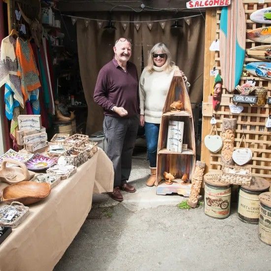 Man and woman stand in front of a craft booth in Salcombe. They smile near wood crafts and a shelf.
