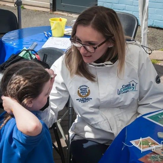 Woman in white jacket leans toward child, talking, at a table covered in blue cloth, outdoors.
