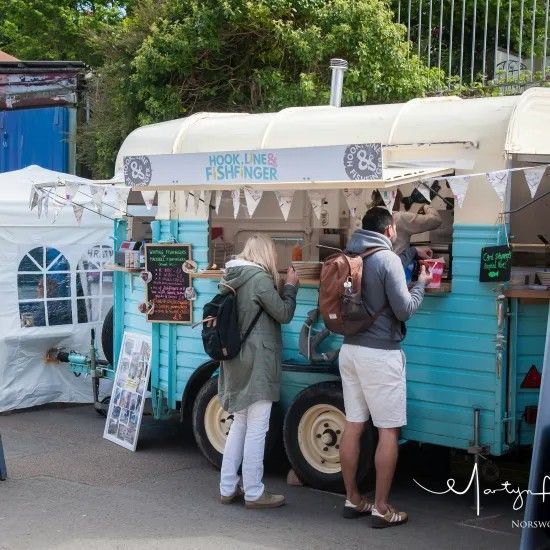 A turquoise food trailer with a queue; two people order food from the window, and a sign says 