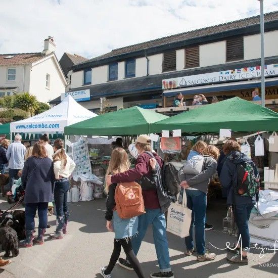 Outdoor market, people browsing stalls under tents and awnings. Buildings in the background.