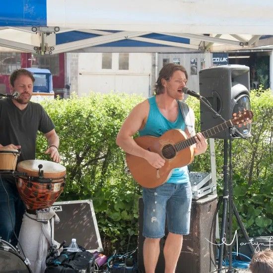 Two musicians perform outdoors: one plays guitar and sings, the other plays a drum.