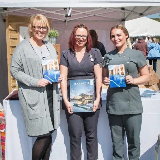 Three women holding brochures at an outdoor event booth; grey outfits, smiles, and sunny setting.
