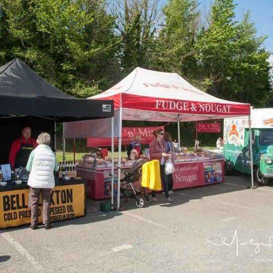 Outdoor market stalls: fudge, nougat, cold-pressed oil, and food truck under tents on a sunny day.