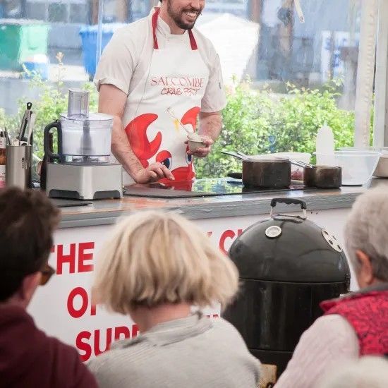 Chef in apron cooking at an outdoor food demonstration. Spectators watch.