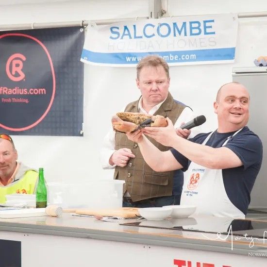 Man holding up food, smiling, with a microphone; another man watches. Demonstration setting.
