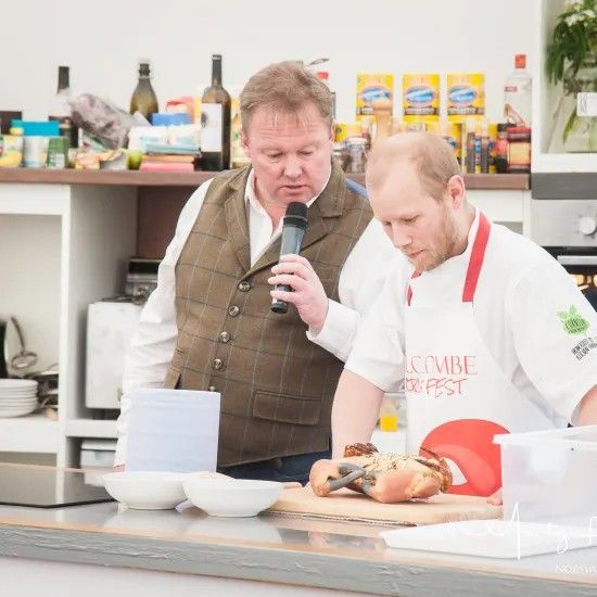 A man with a microphone speaks to a chef who is preparing food at a kitchen counter.