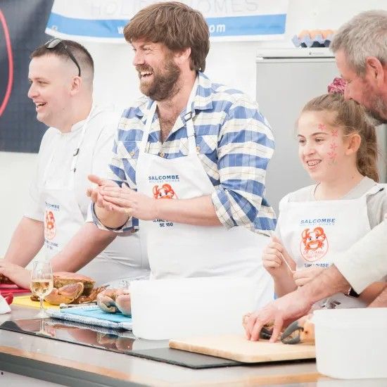 People in aprons smile and clap at a cooking demonstration; food on counter.