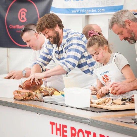 People preparing food at a counter. One man carves meat, others cut ingredients. The setting appears to be a food event.