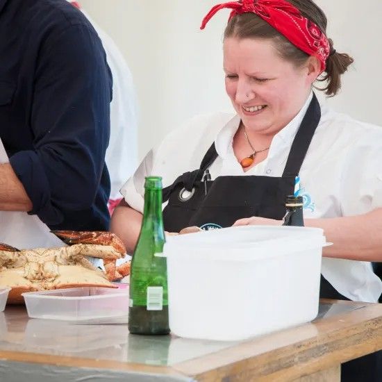 Woman wearing a red bandana and apron smiles while working at a table, with food and a green bottle in view.