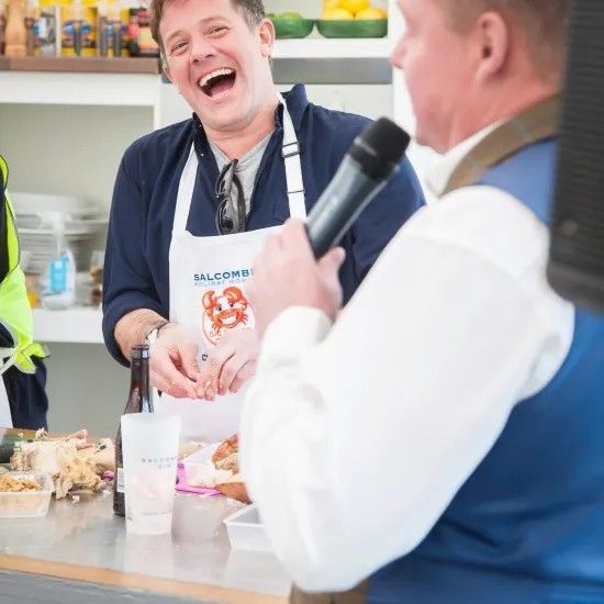 Man laughs while another holds microphone at food event.