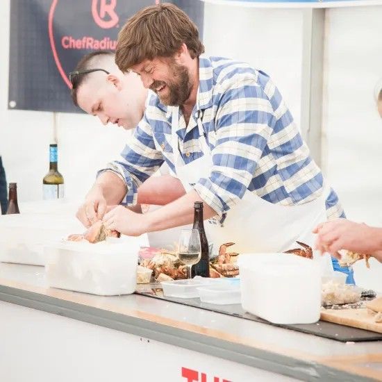 Two men in aprons preparing food at an outdoor event, one wearing blue striped shirt, the other with a shaved head.