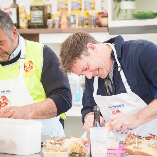 Two men in aprons preparing food at a counter, one laughing, with food containers visible.