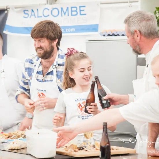 People at a cooking event in Salcombe. A girl holds bottles as others prepare food.