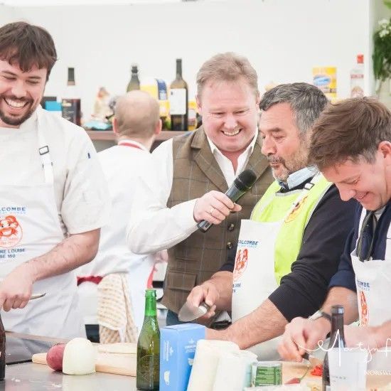 Chefs at a cooking competition, smiling and working. One holds a microphone, a bright event.