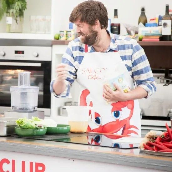 Man in apron cooking in a kitchen, smiling. Includes food processor, bowls, and ingredients.