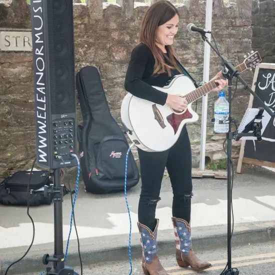 Woman playing white acoustic guitar outdoors, wearing cowboy boots, smiling.