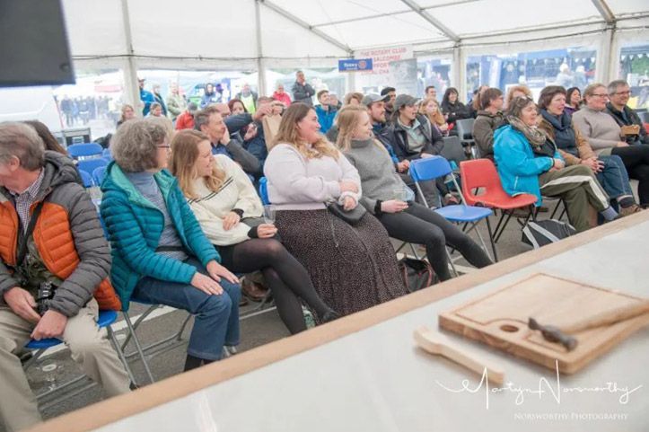 People seated at an event, inside a tent. A wooden cutting board and utensils are in the foreground.