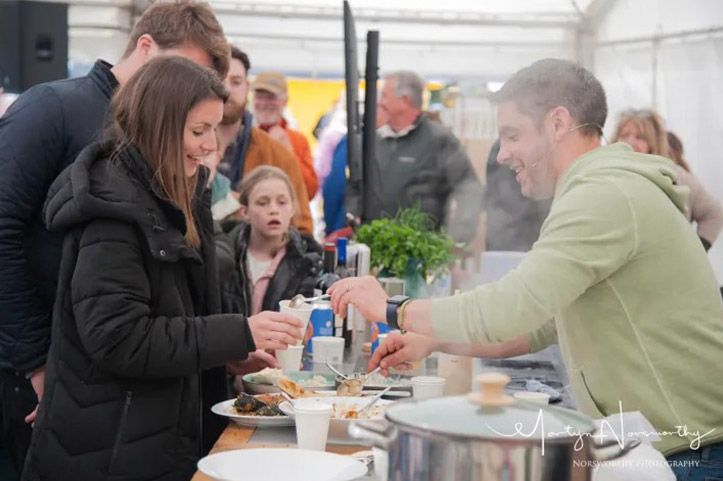People at an outdoor food stall; a man serves food to a woman. Others look on, some smiling.