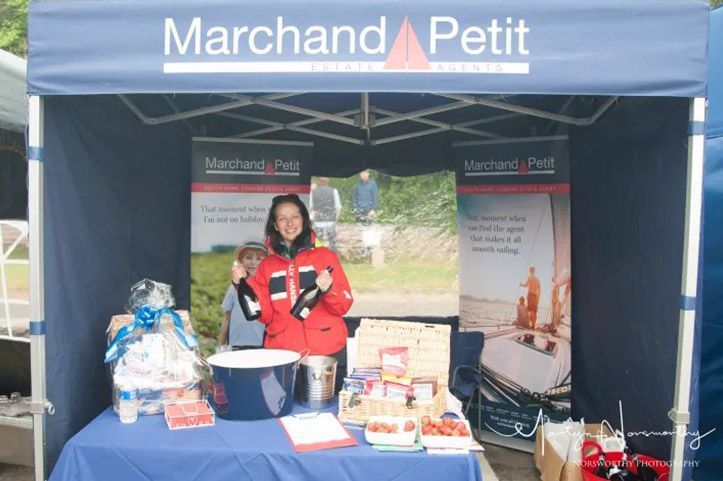 Woman in red jacket at a Marchand Petit stand, holding bottles, smiling. Blue tent, products on table.