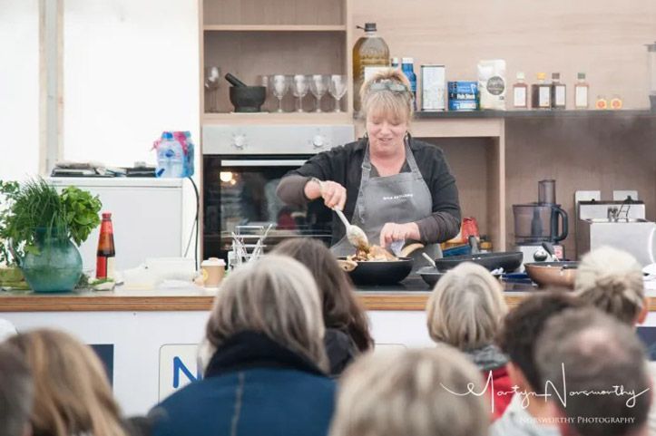 Woman cooking in front of a seated audience. She stirs food in a pan; kitchen setting with ingredients.