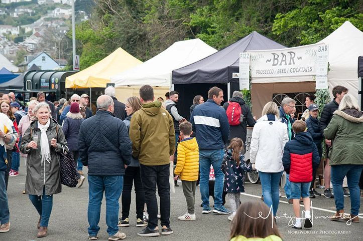Crowd at an outdoor market with tents and bar sign; people walking, dressed in casual wear, under overcast sky.