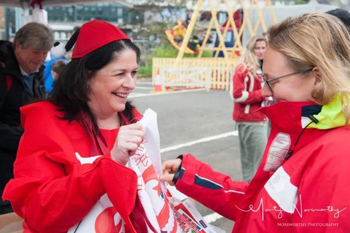 Two people in red jackets exchanging a bag outdoors; one smiles, wearing a fez-like hat.