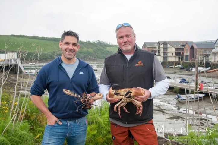 Two men holding a lobster and a crab near a harbor. One man smiles, the other looks forward.