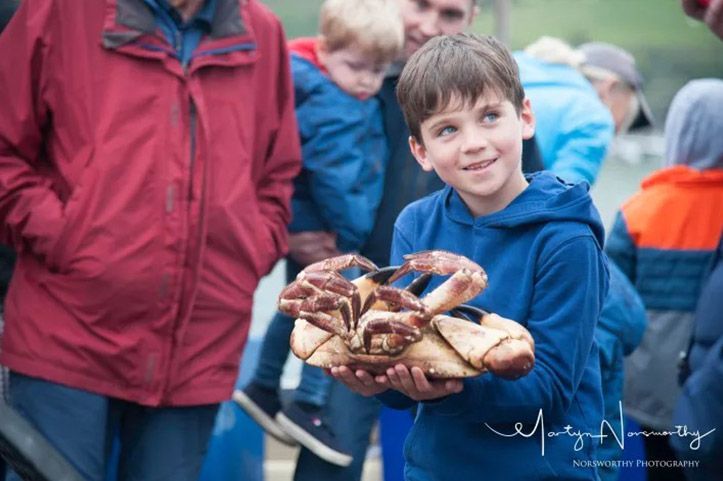 Boy in blue holds large cooked crabs, smiling; other people and water in background.