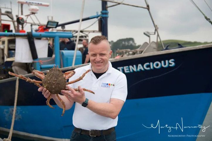 Man holding a large crab in front of a blue fishing boat labeled 