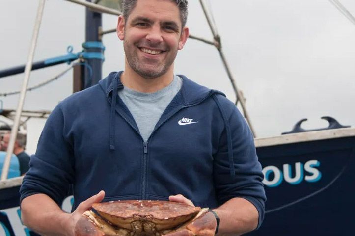 Man holding a large crab, smiling, in front of a fishing boat. He wears a blue jacket.