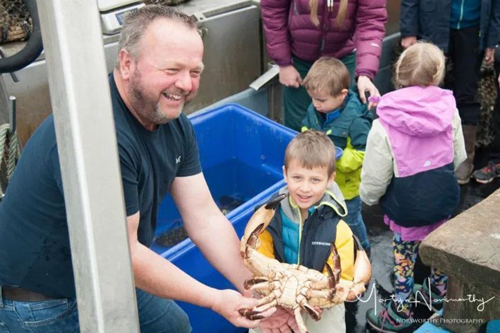 Man and young boy holding large crab, smiling, on a boat with children and blue containers.