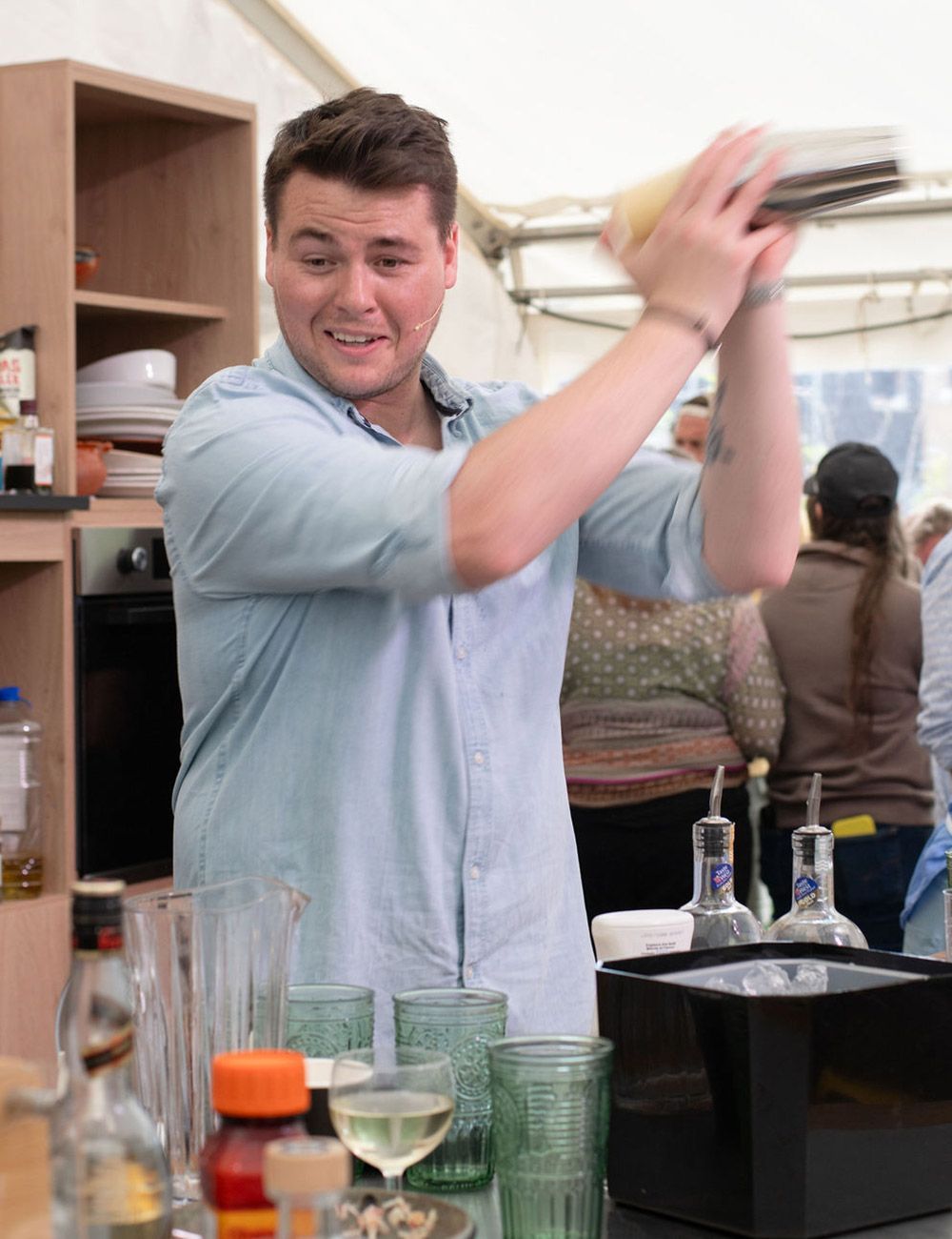 Bartender shaking a cocktail at a bar. He wears a blue shirt, smiling, with glassware and bottles nearby.