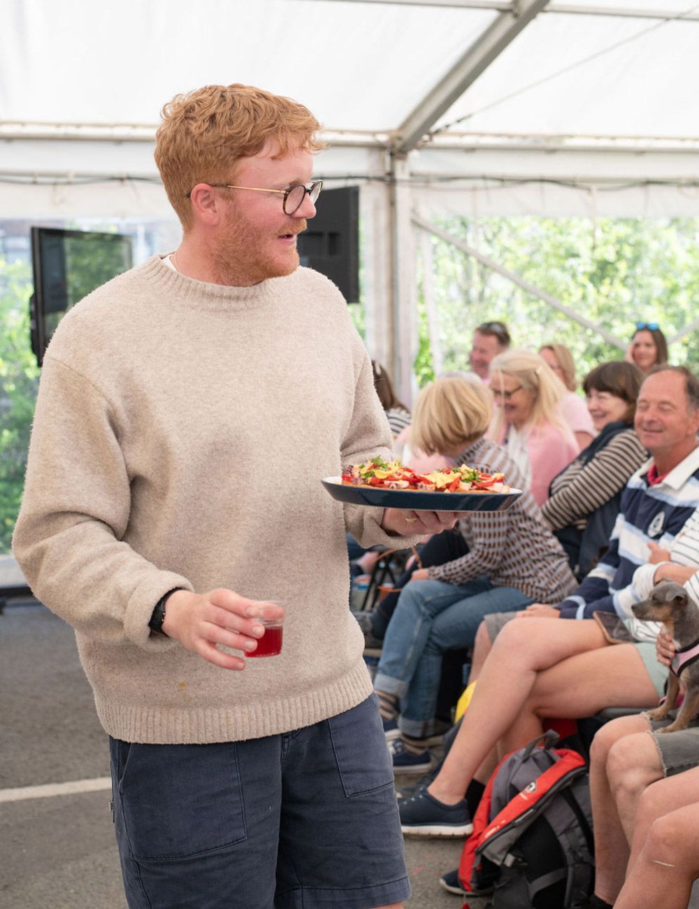 Man with red hair and glasses holding a tray of food and a drink, smiling, inside a tent with people seated behind him.