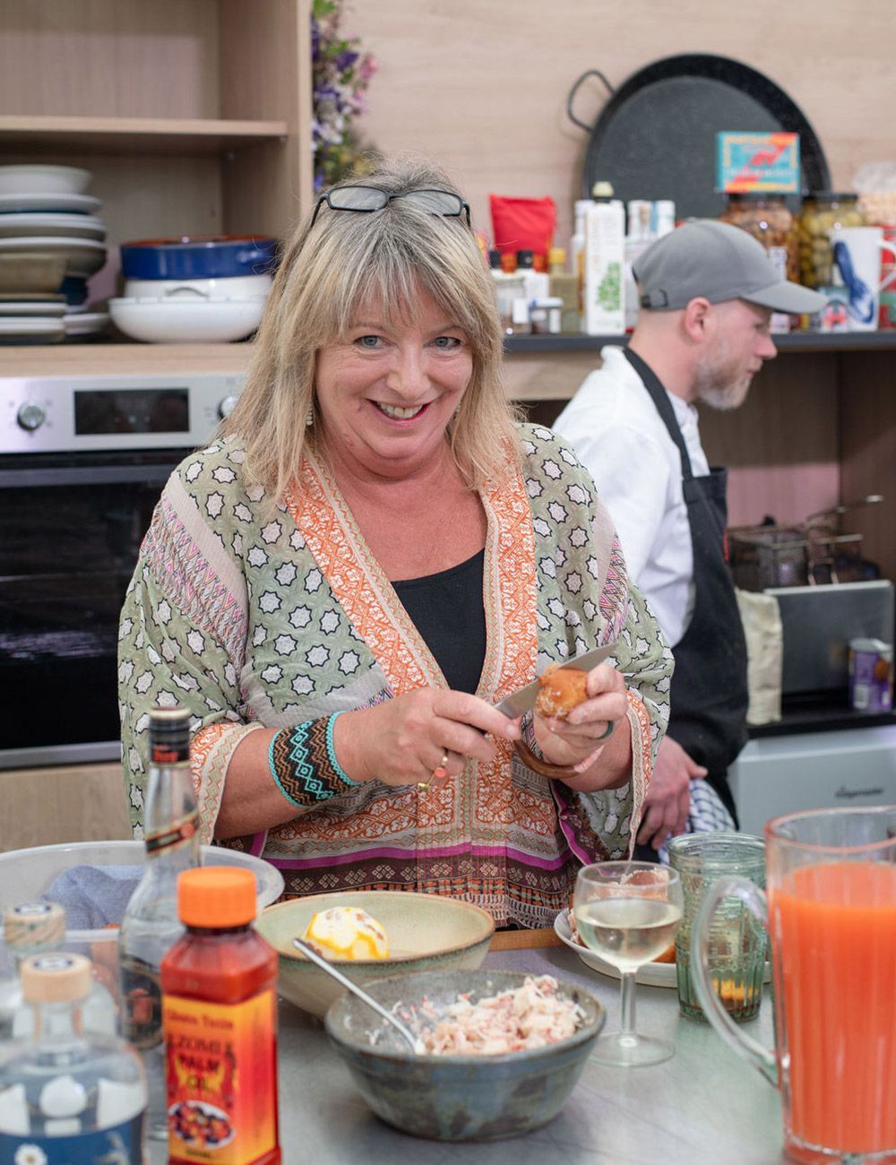 Woman in kitchen, smiling, cutting food. Behind her, a person in a chef's apron. Bowls and bottles on a counter.
