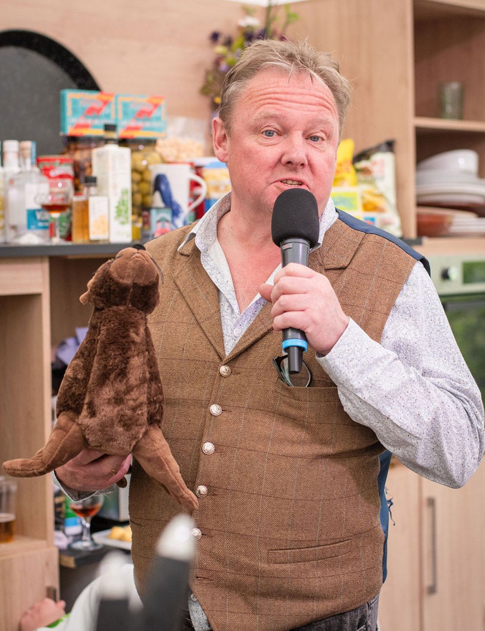 Man holding a microphone and a brown stuffed animal, speaking. He wears a vest and stands in a kitchen.