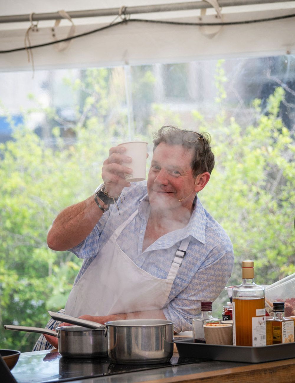 Man in white apron and blue shirt, smiles while holding a steaming cup near pots on a stove.