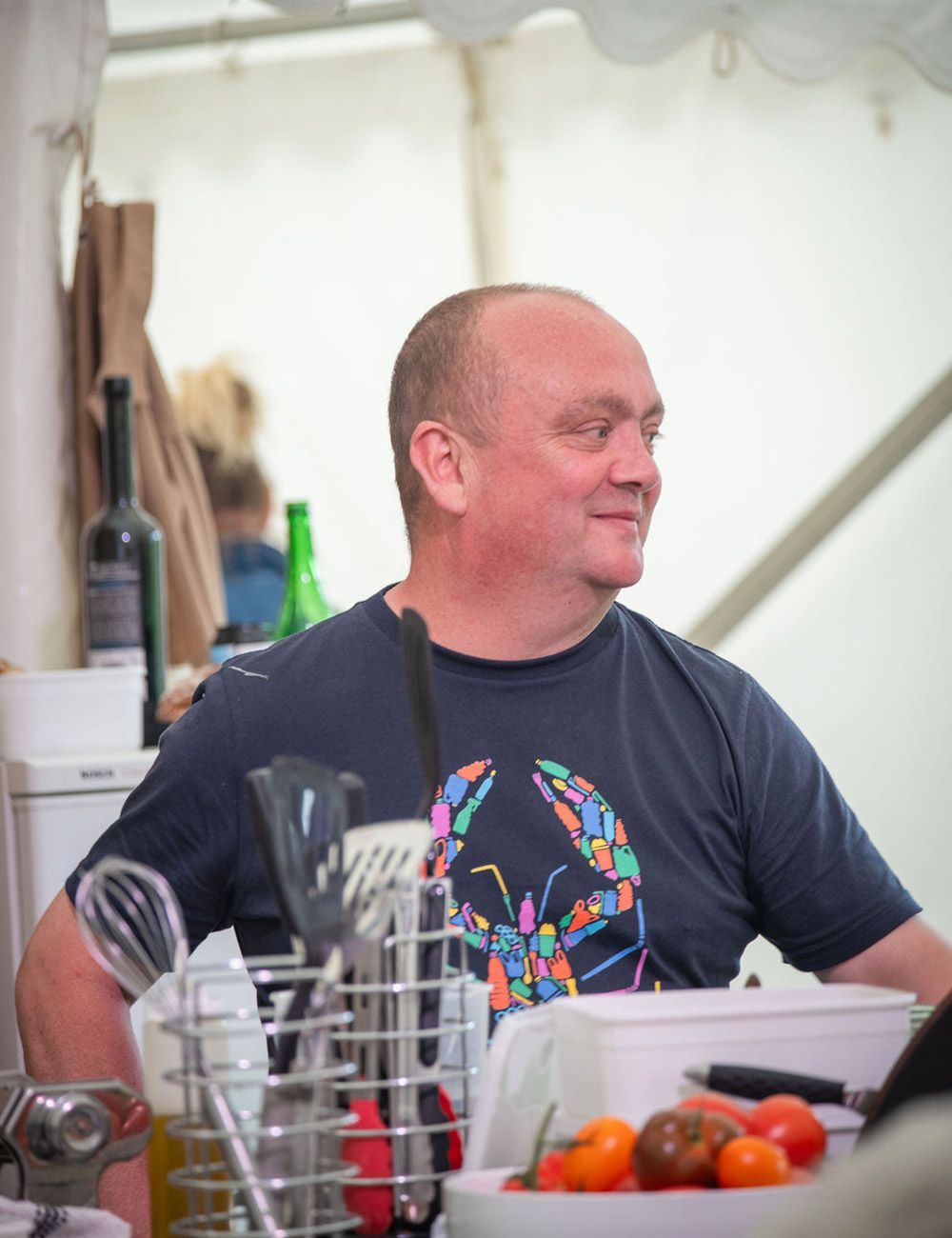 Man wearing a navy shirt with a colorful lobster graphic smiles, standing behind a counter with cooking utensils and produce.