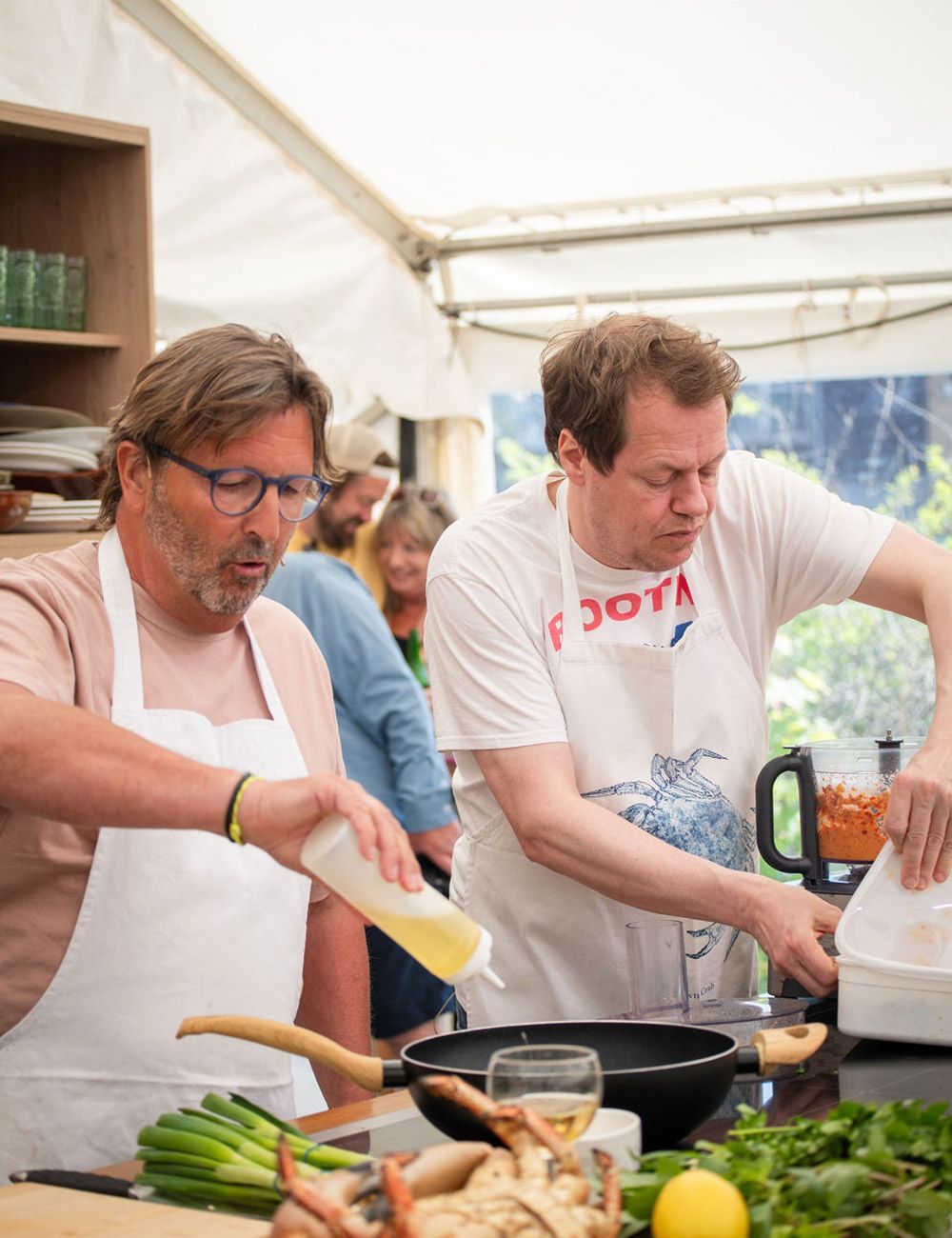 Two men cooking in a tent, one pouring oil, the other using a food processor.