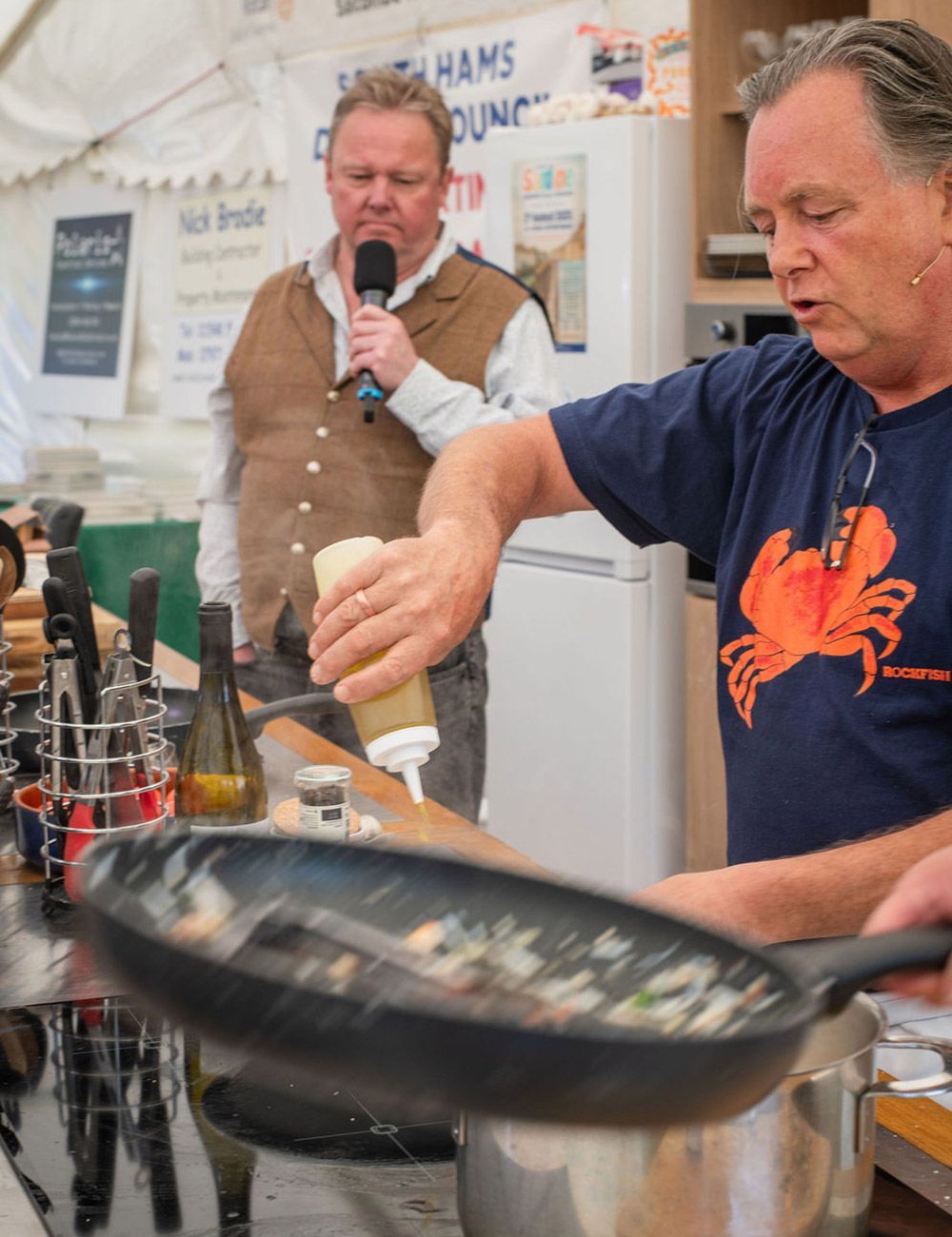 Man cooking food in a pan, pouring sauce. Another man holding a microphone in the background.