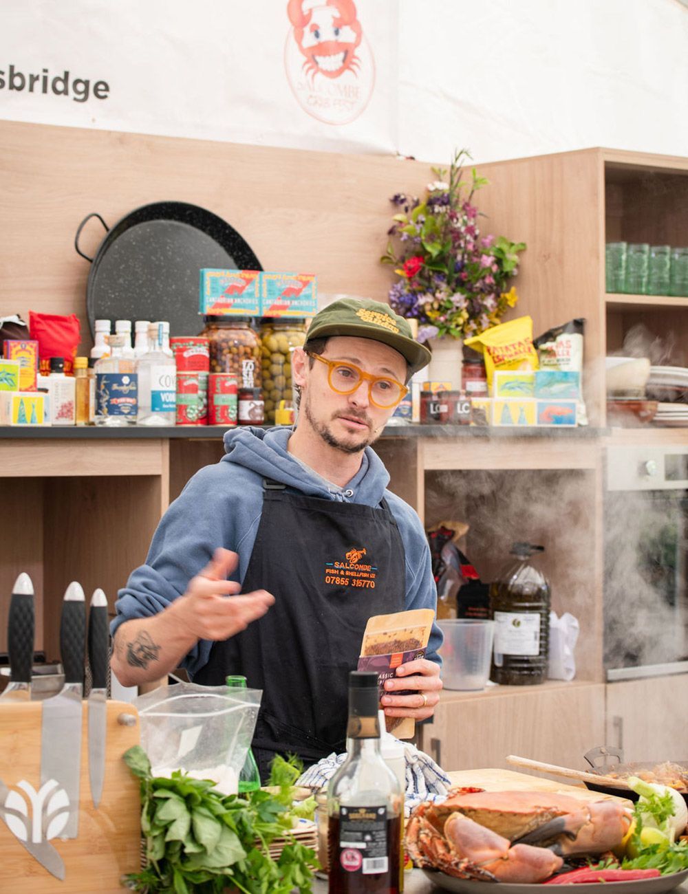 Chef in apron gestures toward food, cooking in a brightly lit kitchen with ingredients visible.