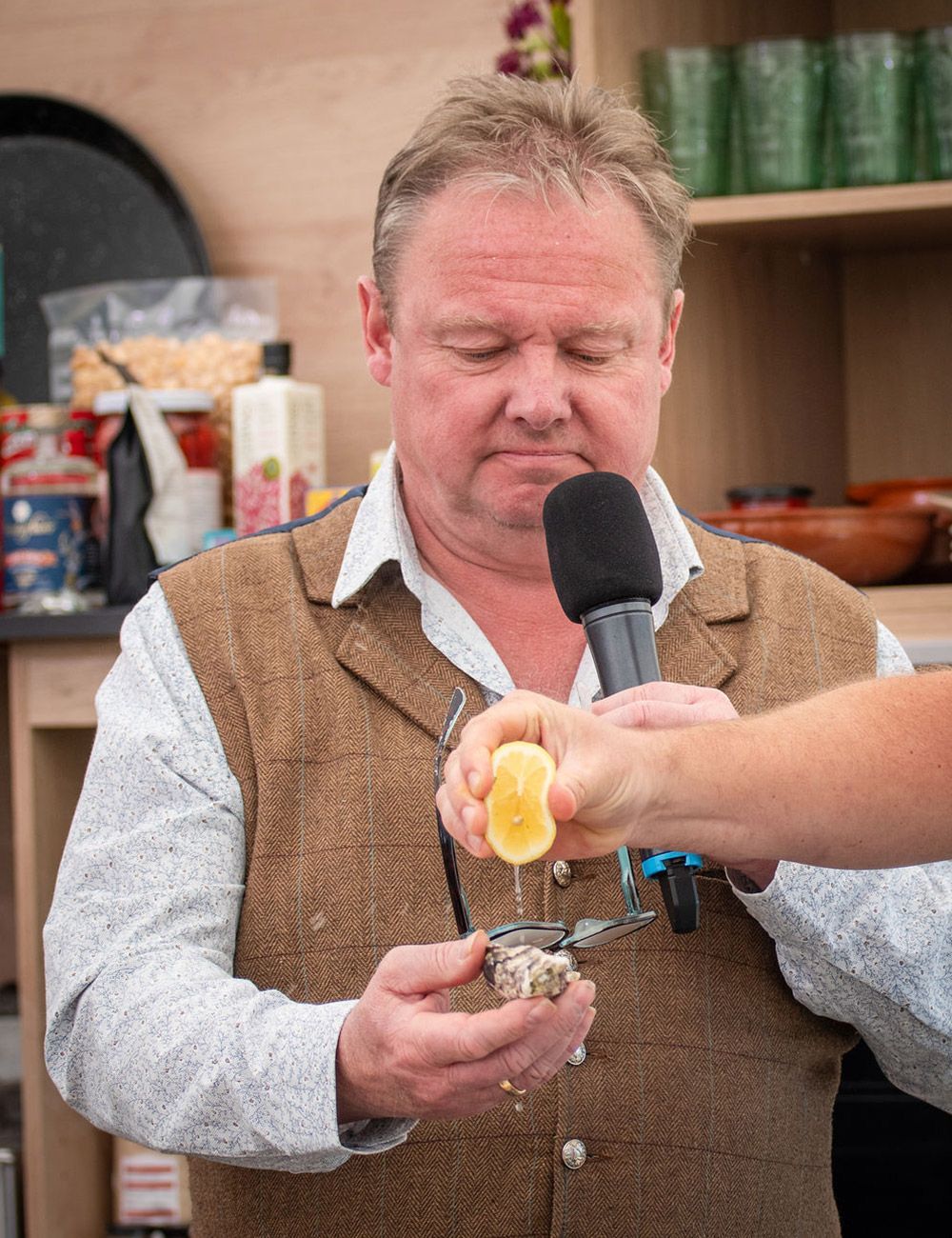 Man squeezing lemon juice onto an oyster, holding a microphone. Outdoors, preparing food.