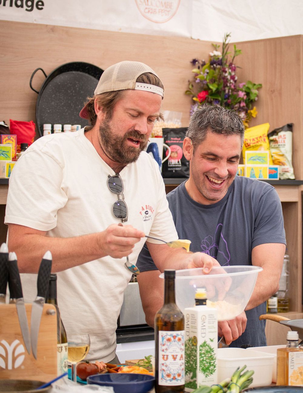 Two men cooking outdoors, one stirring, the other laughing. Food and ingredients on counter.