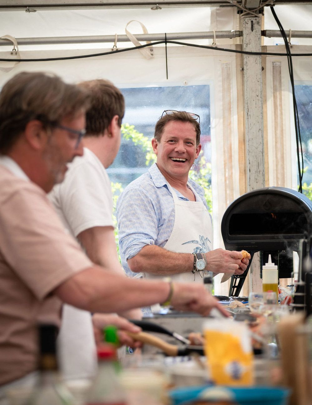 Three people cooking together outdoors; one man smiling, wearing an apron, near a grill and a white tent.