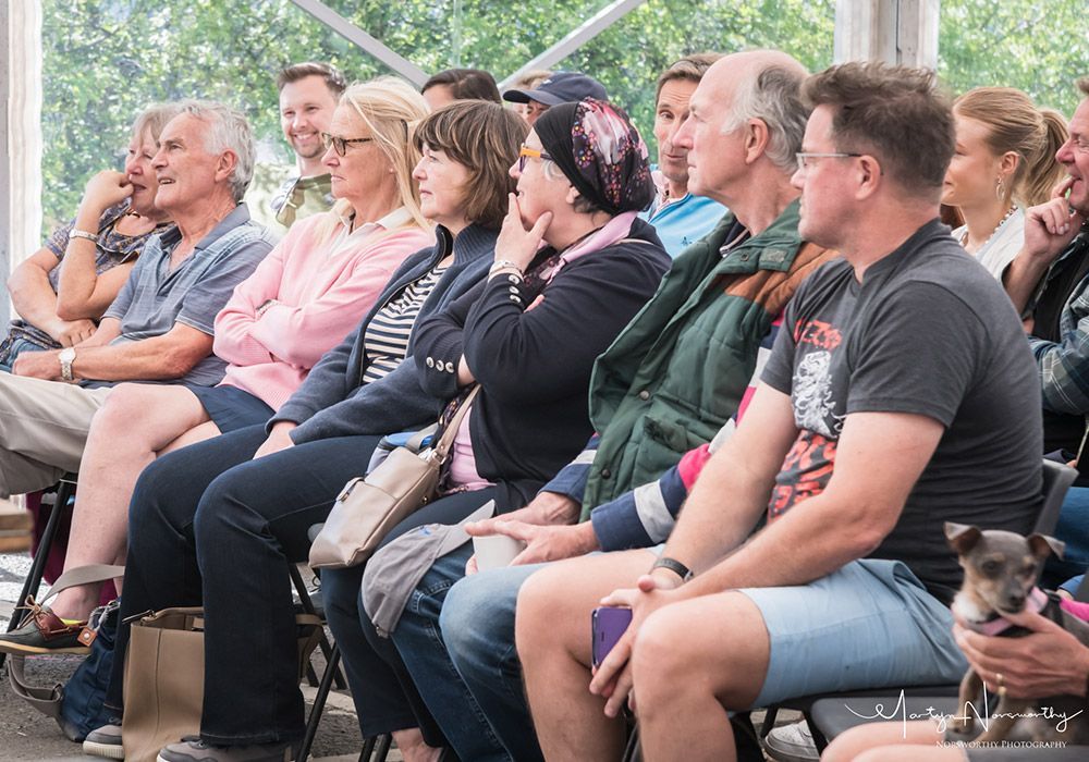 Group of people seated, watching something outdoors. Some have contemplative expressions, and one holds a small dog.