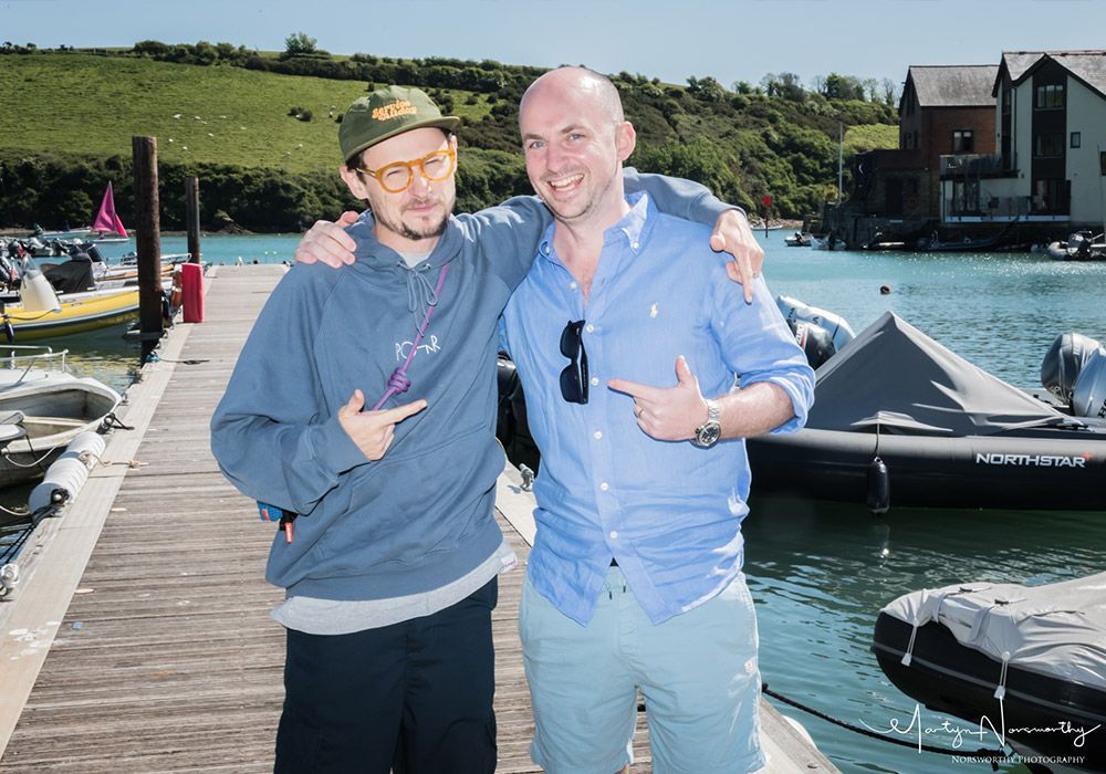 Two men on a dock, arm around each other, pointing. Blue water, boats, and green hillside in background. Sunny day.